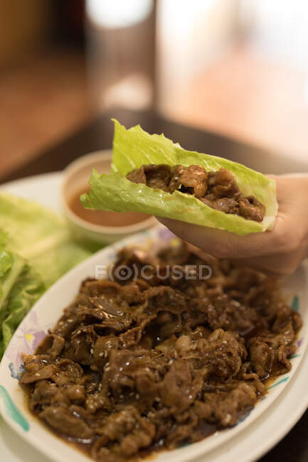 Рука держит приготовленное аппетитное мясо, завернутое в свежий здоровый салат над тарелкой на деревянном столе в кафе — стоковое фото