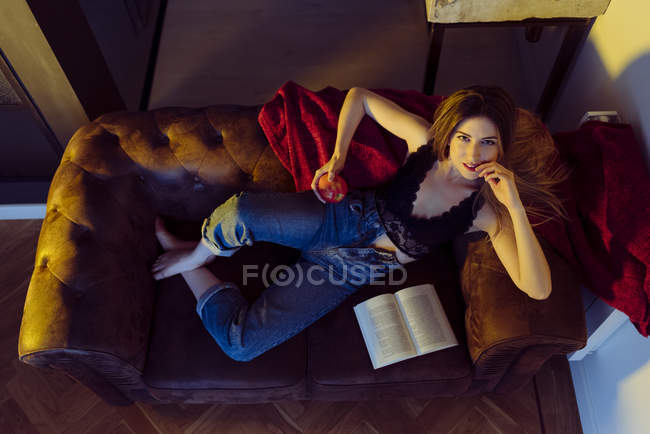 Jeune femme sexy assise sur un canapé avec pomme et livre — Photo de stock