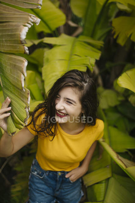 Задумчивая молодая женщина в желтой футболке поддерживает голову и смотрит в сторону, сидя рядом с экзотическим кустарником в саду — стоковое фото