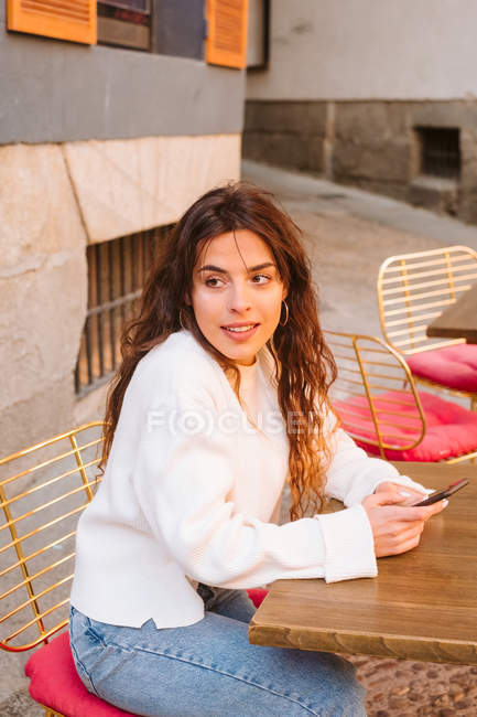 Молодая женщина в повседневной одежде сидит за столом в открытом кафе со смартфоном на городской улице — стоковое фото