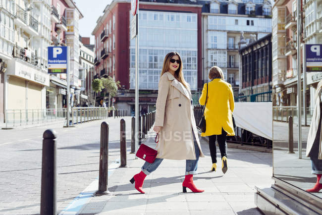Young happy trendy woman in trench-coat and sunglasses walking in city — Stock Photo