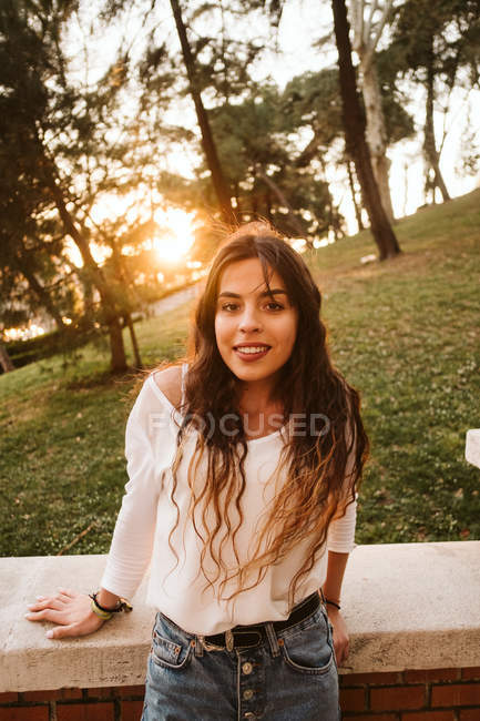 Pretty young woman in casual outfit smiling and looking at camera while leaning on border on sunny day in park — Stock Photo