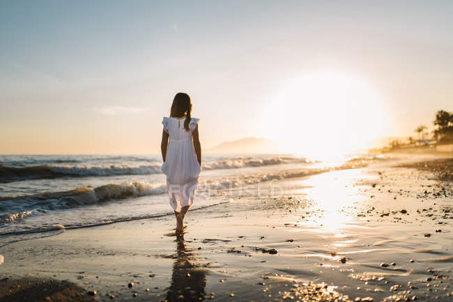 Little girl in white dress walking on seashore on background of sunset ...