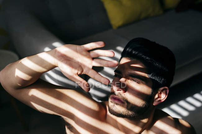 D'en haut jeune homme beau nu avec coiffure élégante debout près du canapé et couvrant la lumière du soleil avec la main — Photo de stock