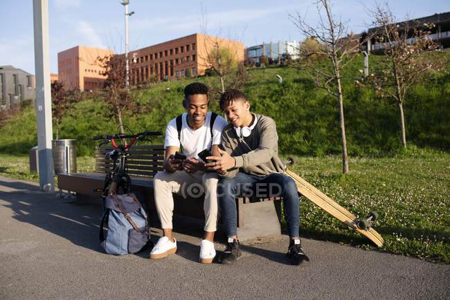 Jóvenes hombres negros sentados en el banco con smartphone - foto de stock