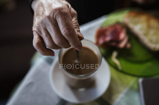 Immagine ritagliata di donna che tiene cucchiaio in tazza durante la miscelazione di caffè a colazione — Foto stock