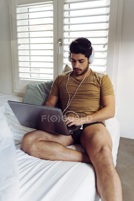 Jovem homem bem sucedido concentrado em camisa casual e fones de ouvido sentado na cama usando um laptop — Fotografia de Stock