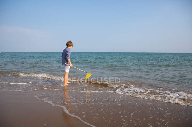Side view of long hair cute boy in summer wear standing in water with fishing net on sea shore in sunny day — Stock Photo