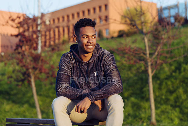 Joven hombre negro sentado en el banco con monopatín - foto de stock