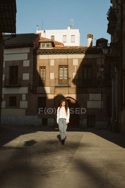 Trendy young woman in casual outfit walking on pavement of aged town on sunny day — Stock Photo