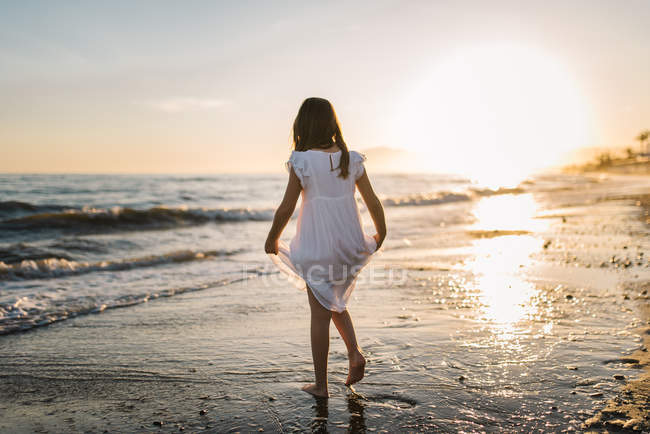 Kleines Mädchen in weißem Kleid, das im Wasser am Strand vor dem Hintergrund der Sonne spaziert — Stockfoto