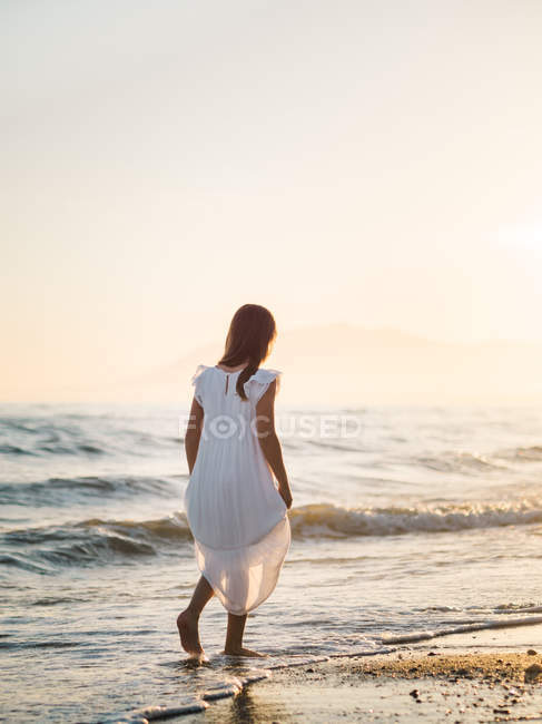 Kleines Mädchen in weißem Kleid, das am Ufer des Meeres im Hintergrund des Sonnenuntergangs spaziert — Stockfoto