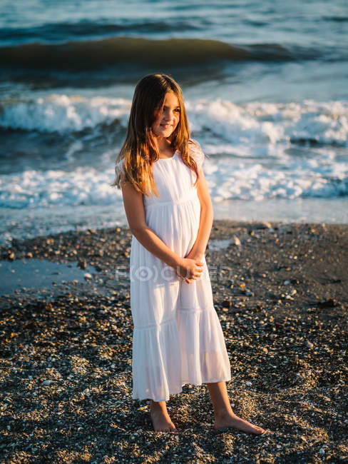 Portrait of charming little girl in white dress standing on beach — Stock Photo