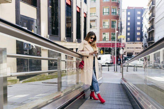 Молода жінка спирається на перила ескалатора зі смартфоном у місті — стокове фото