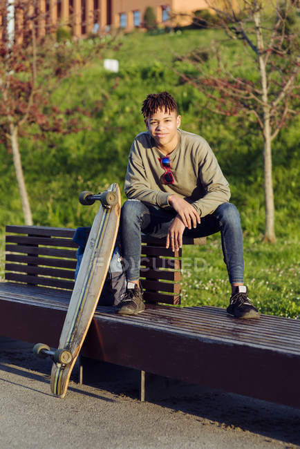 Молодой черный мужчина сидит на скамейке со скейтбордом — стоковое фото