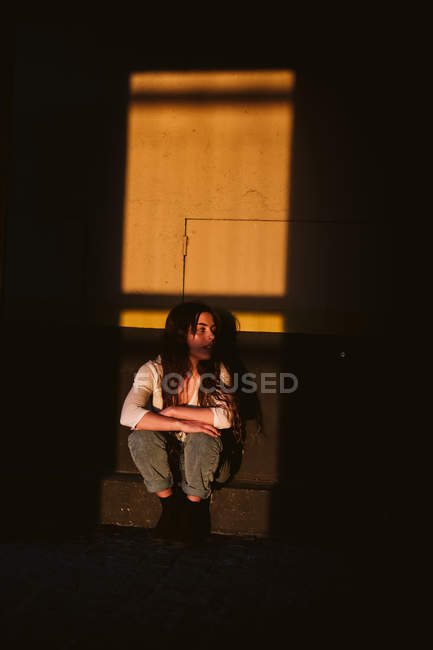 Young female in casual outfit sitting on building wall in spot of sunlight — Stock Photo