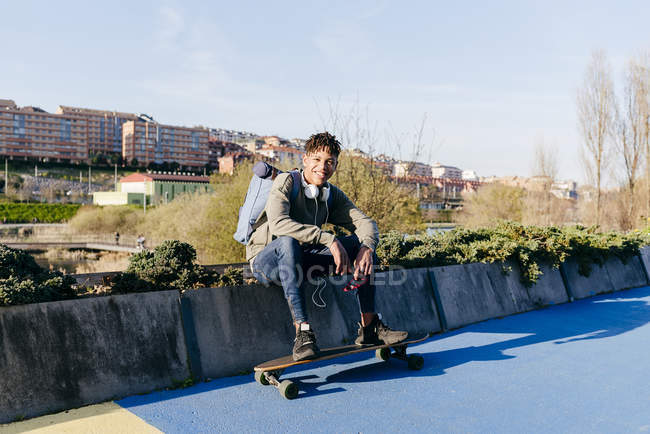 Молодой черный радостный красивый мужчина с афроволосами в повседневной одежде и наушниках, сидящий со скейтбордом и расслабляющий — стоковое фото