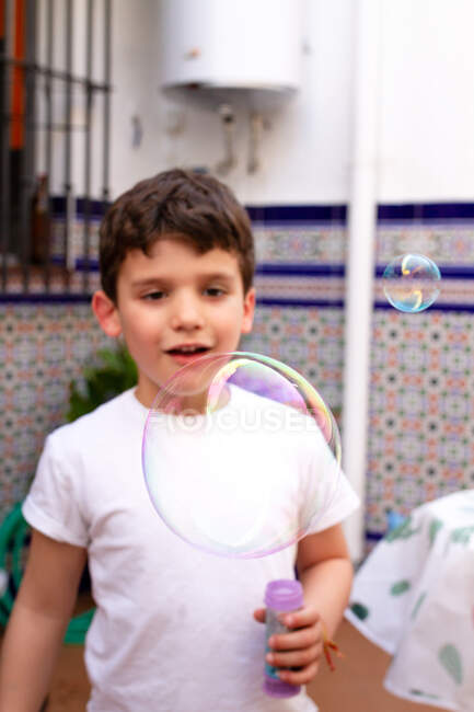 Menino de camiseta branca jogando bolhas de sabão enquanto estava no terraço em casa — Fotografia de Stock