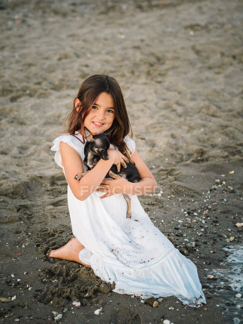 Portrait de charmante fille en robe blanche tenant un petit chien assis sur du sable — Photo de stock