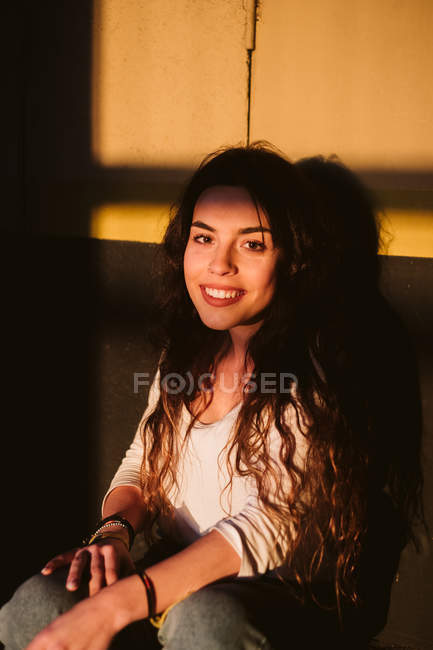 Молодая женщина в повседневной одежде улыбается, сидя перед стеной здания в месте солнечного света — стоковое фото