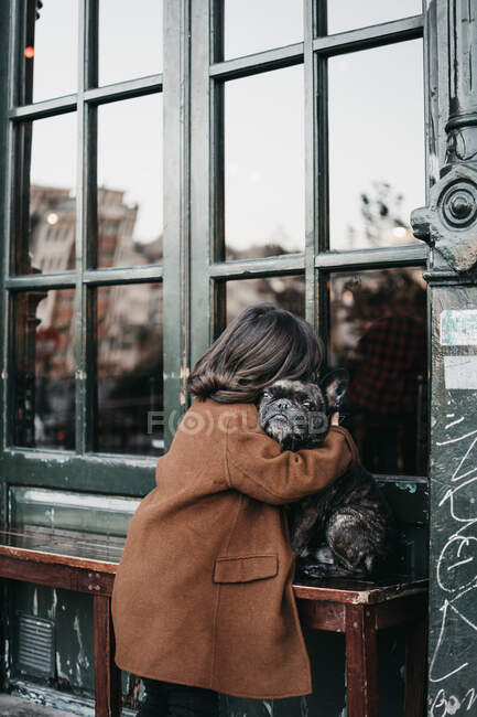 Side view of young female in casual warm coat hugging adult sad beautiful dog on table on stone beach — Stock Photo