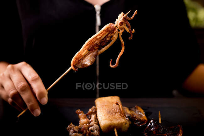 Hembra comiendo apetitoso brocheta de calamar caliente a la parrilla sobre el plato con brochetas de carne y verduras en la mesa de madera en la cafetería - foto de stock