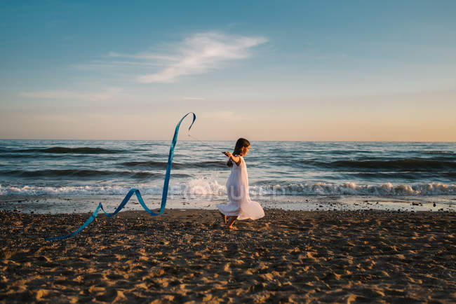 Little girl in white dress running along seashore and holding long blue band on background of evening sky — Stock Photo