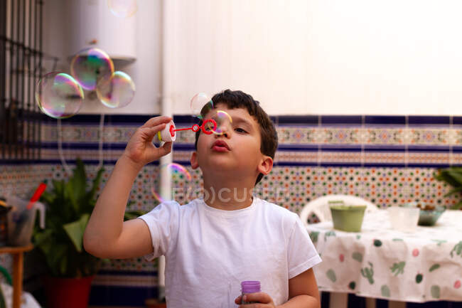 Pequeño niño en camiseta blanca soplando burbujas de jabón mientras está de pie en la terraza en casa - foto de stock