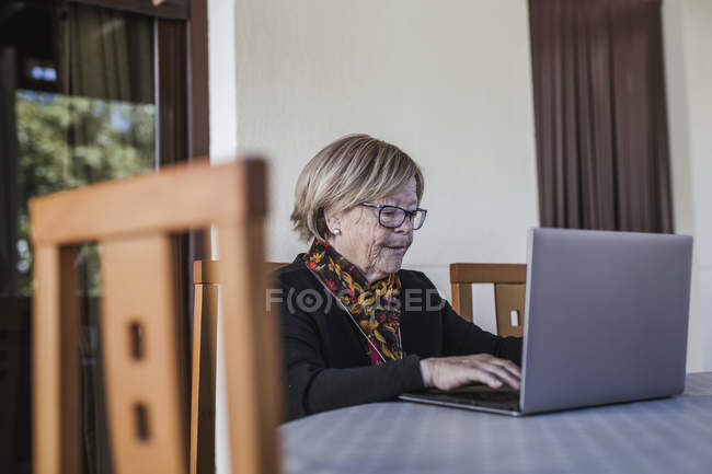 Літня жінка в окулярах переглядає ноутбук, сидячи за столом у затишному будинку — стокове фото