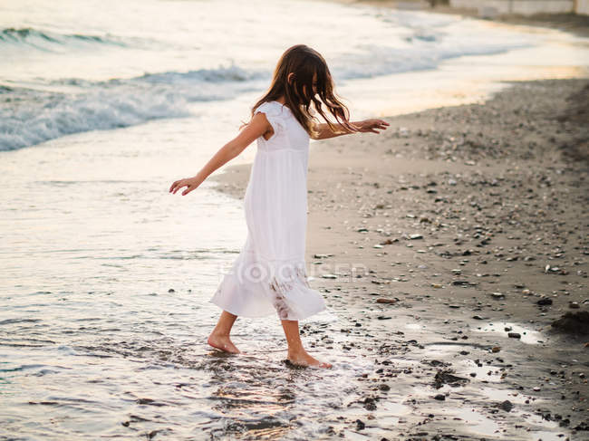 Маленькая девочка в белом платье, гуляющая в воде по пляжу — стоковое фото
