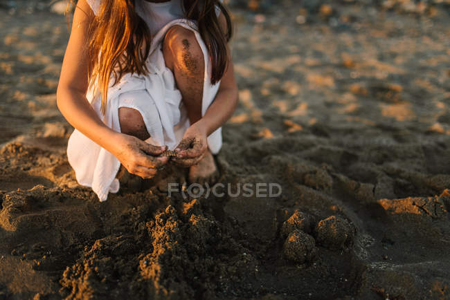 Nahaufnahme eines weiblichen Kindes im weißen Kleid, das am Strand im Sonnenlicht mit Sand spielt — Stockfoto