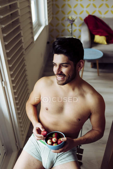 Vista lateral de un joven alegre y sexy hombre exitoso en ropa interior sentado en una silla de madera y comiendo fresa cerca del balcón - foto de stock