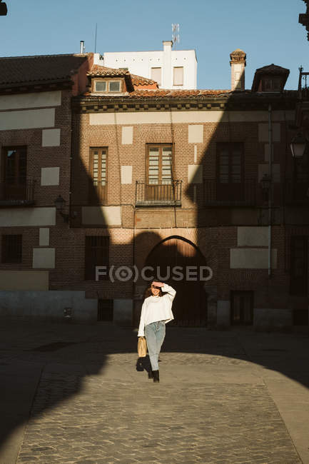 Mujer joven de moda en traje casual caminando en el pavimento de la ciudad envejecida en el día soleado - foto de stock