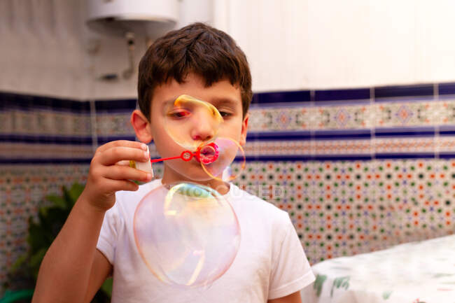 Kleiner Junge im weißen T-Shirt pustet Seifenblasen, während er zu Hause auf der Terrasse steht — Stockfoto