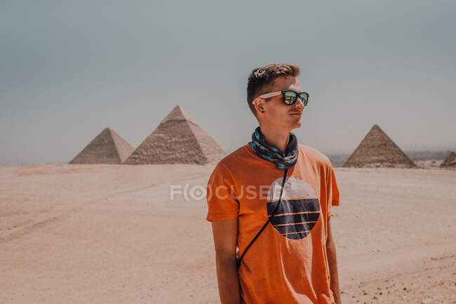 Уверенный мужчина путешественник в солнцезащитных очках глядя в сторону, стоя в пустыне против известных Великих пирамид в Каире, Египет — стоковое фото