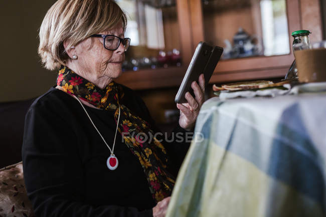 Seniorin schaut und berührt Smartphone-Bildschirm, während sie auf Sofa im Wohnzimmer sitzt — Stockfoto
