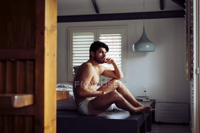 Young pensive successful sexy man with stylish hairstyle in grey briefs lying on bed at home and looking away — Stock Photo