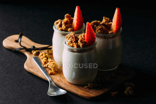 Glasses of cold tasty milk and delicious granola with strawberries on wooden board on black background — Stock Photo