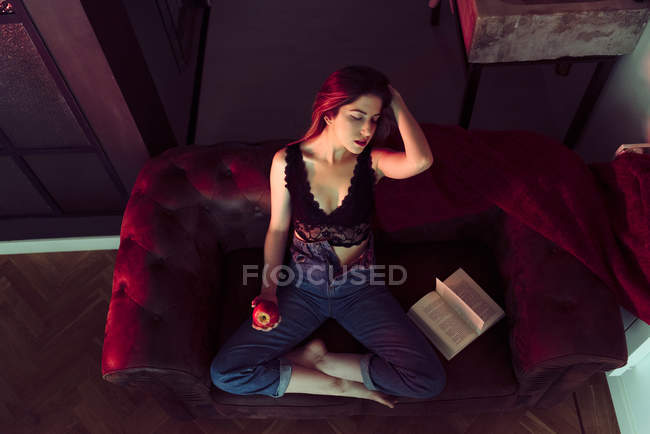 Jovem mulher sexy sentado no sofá com maçã e livro — Fotografia de Stock