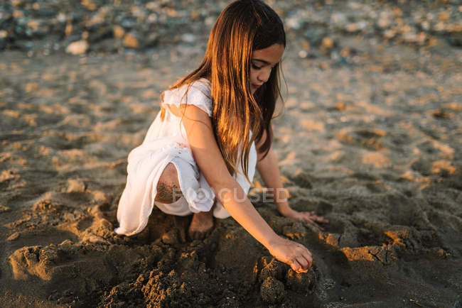 Lindo niño femenino pensativo en vestido blanco jugando con arena en la playa a la luz del sol - foto de stock