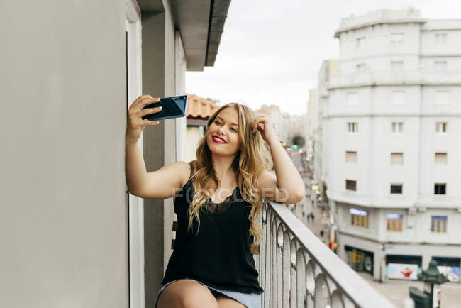 Giovane donna bella felice con i capelli biondi in abito casual seduto sul balcone e prendere selfie — Foto stock