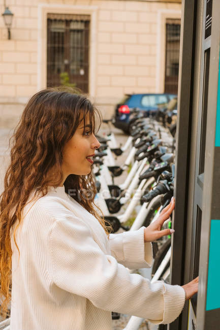 Jeune femme souriante en tenue décontractée en utilisant kiosque sur la station de partage de vélos sur la rue de la ville — Photo de stock