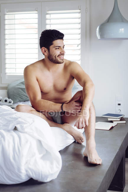 Молодой улыбающийся успешный сексуальный мужчина со стильной прической в серых трусах, лежащих на кровати дома и смотрящих в сторону — стоковое фото
