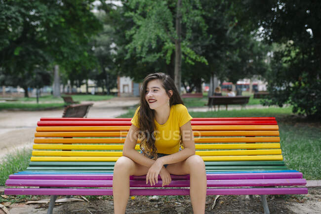 Hübsche junge Frau in lässigem Outfit lächelt fröhlich und schaut weg, während sie auf der Regenbogenbank im Park sitzt — Stockfoto