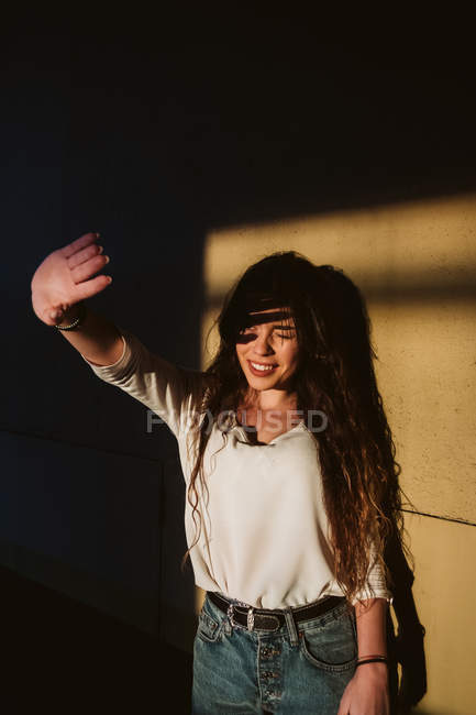 Jeune femme en tenue décontractée bloquant le soleil avec la main et souriant contre le mur — Photo de stock