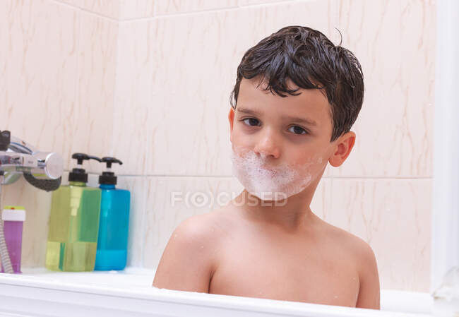 Adorable kid looking at camera with mouth covered in foam while sitting in the bath in bathroom — Stock Photo