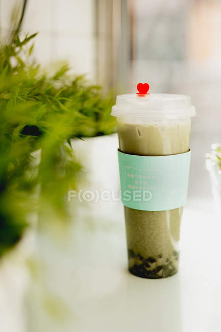 Tazza di plastica con cuore di gustoso tè matcha caldo sul tavolo vicino a piante in vaso nel ristorante asiatico . — Foto stock