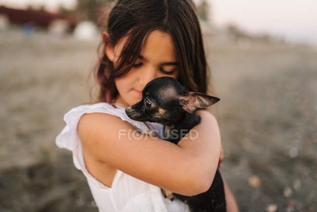 Portrait of charming female child in white dress holding little dog on beach — Stock Photo