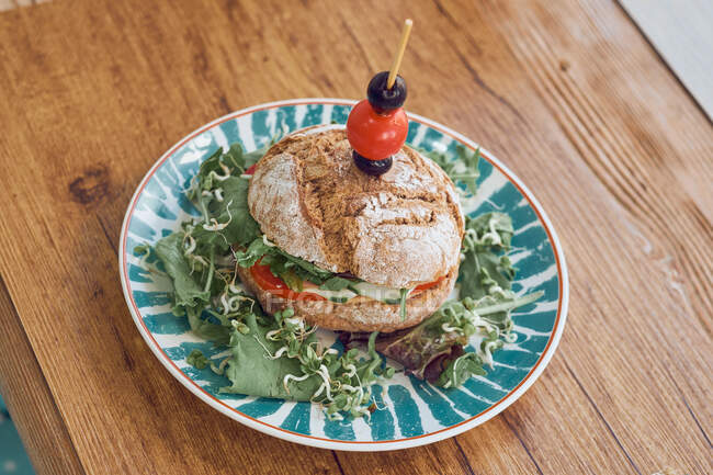 Delicioso sándwich vegetariano con tomates, cebolla, lechuga, aceitunas y tomate cherry en plato brillante sobre mesa de madera en restaurante - foto de stock