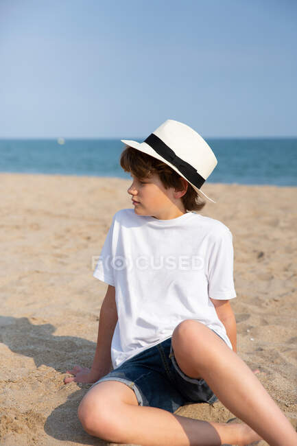 Милый мальчик в белой футболке и шляпе, сидящий на песчаном пляже в летний день — стоковое фото